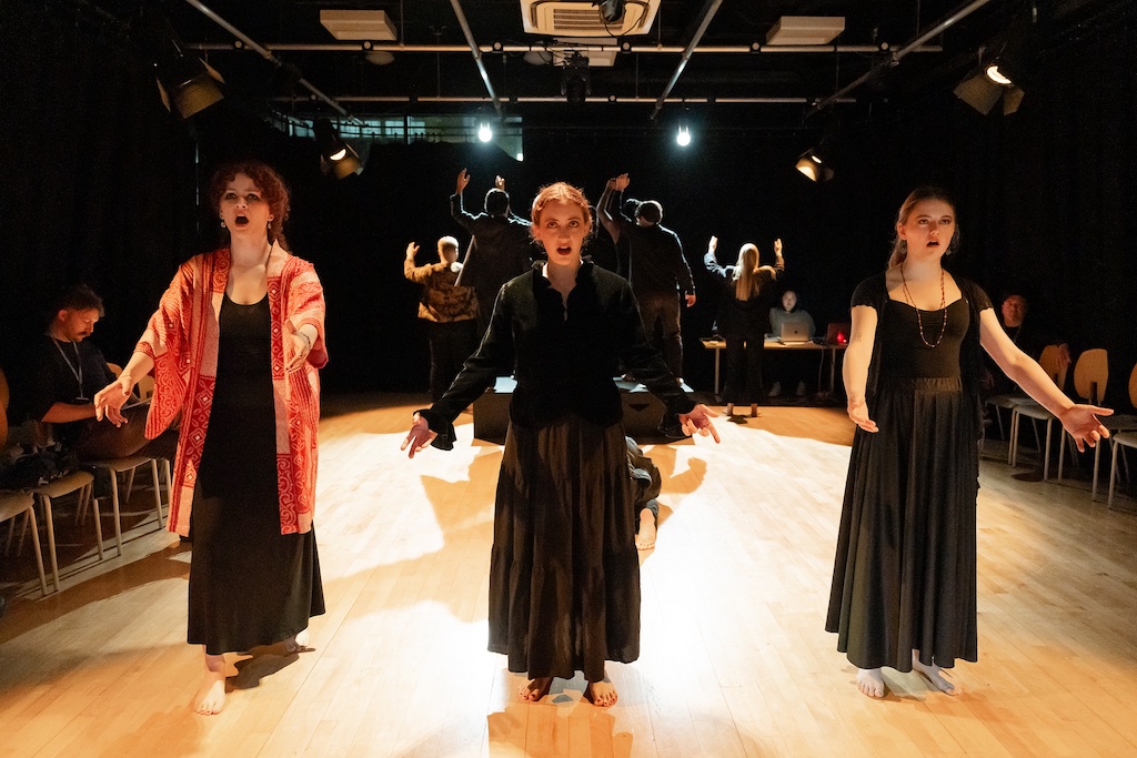 Three actors wearing long black dresses stand with their arms outstretched on stage in mid-performance. The actor on the left is wearing a patterned light red coat over her dress.